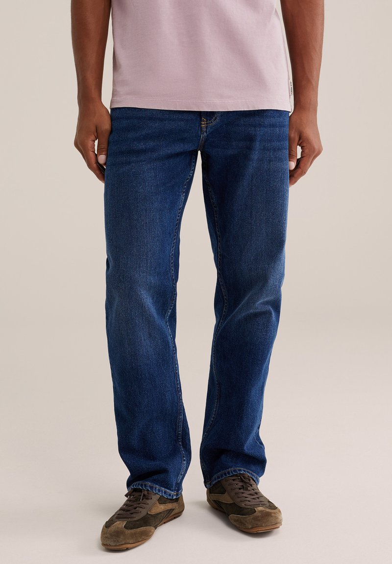 Homme portant un jean bleu droit, des baskets en daim marron et un t-shirt rose clair, debout sur un fond neutre.