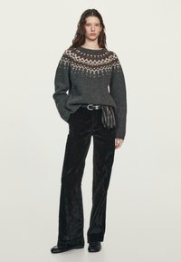 Grauer Strickpullover mit gemustertem Passe, kombiniert mit schwarzen Cord- Schlaghosen. Das Modell steht mit den Händen in den Taschen und trägt Lederhandschuhe.