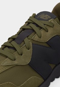 Εγγύς πλάνο αθλητικού παπουτσιού New Balance σε ελιά πράσινο και μαύρο χρώμα με υφασμάτινη υφή, λεπτομέρειες από σουέτ και κορδόνια σε χρώμα ελιάς δεμένα πάνω στη γλώσσα.