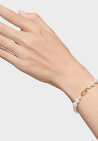 Mano che indossa un braccialetto di perle bianche con chiusura dorata su uno sfondo grigio chiaro uniforme.