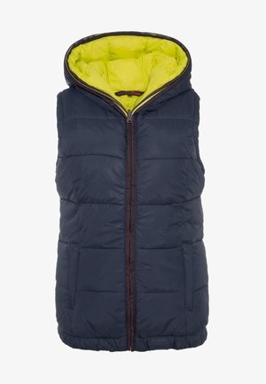 Femme portant un gilet matelassé bleu marine avec une capuche jaune vif, un col roulé bleu marine, un jean clair et des bottines à talons marron, debout.