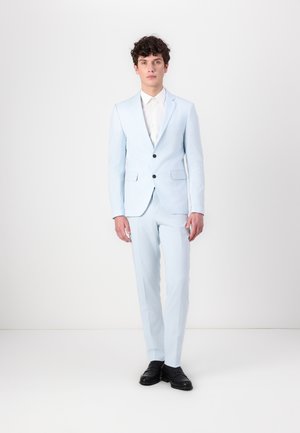 Lyseblå dress laget av glatt stoff, med slank passform, to frontlommer og to sorte knapper. Sammen med en hvit skjorte og svarte sko.