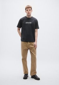 Melna kokvilnas T-krekls ar baltu logo tekstu, kopā ar gaiši brūnām biksēm un melniem apaviem. Vienkāršs dizains un regulārs piegriezums.