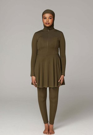 Vrouw staat blootsvoets en draagt een olijfgroene badpak met lange mouwen, een rok en een aangehechte hoofdbedekking.