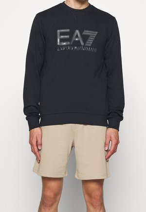 Persona con felpa nera a maniche lunghe EA7 Emporio Armani e pantaloncini beige, in piedi davanti a uno sfondo bianco uniforme.