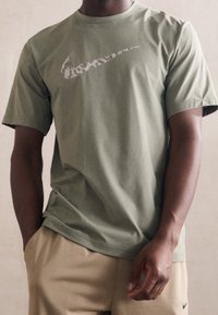 Oliwkowa bawełniana koszulka z krótkim rękawem z białym logo Nike nadrukowanym z przodu. Materiał wydaje się miękki i gładki.