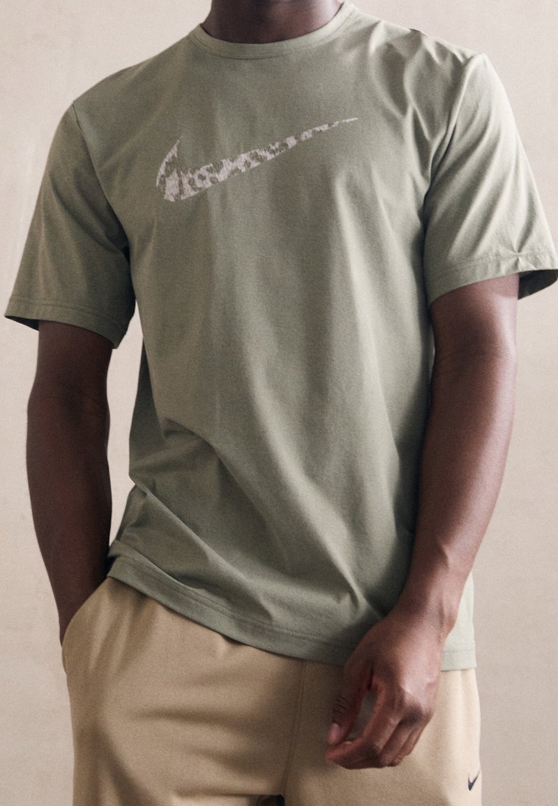Oliwkowa bawełniana koszulka z krótkim rękawem z białym logo Nike nadrukowanym z przodu. Materiał wydaje się miękki i gładki.