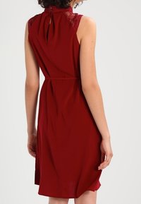 Robe bordeaux à encolure haute, avec des détails en dentelle aux épaules et une ouverture en trou de clé au dos. Tissu fluide avec une taille nouée.