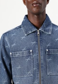 Denim jacket in light blue with a zip closure, pointed collar, two chest pockets, and a subtle star pattern throughout.