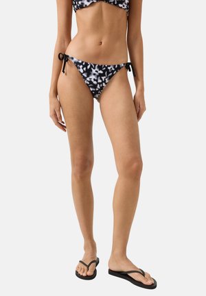 Kvinde iført sort-hvide mønstrede bikinitrusser med bindebånd i siden og sorte flipflops, stående mod en ensfarvet baggrund.