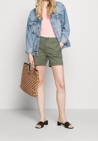 Veste en denim, T-shirt rose, shorts vert olive, sandales à enfiler noires et un sac fourre-tout coloré à motifs avec des poignées.