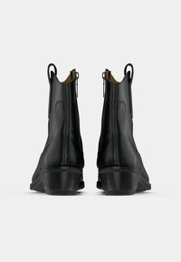 O pereche de bocanci din piele neagră la gleznă, cu toc mic, fermoare laterale și bucle de prindere în spate, prezentată din spate pe un fundal alb.