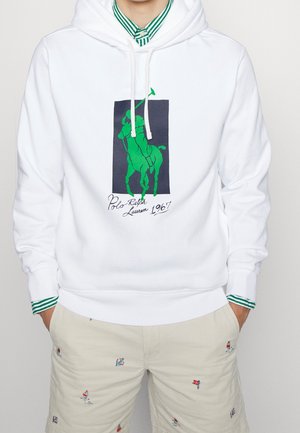 Mann trägt weißen Hoodie mit grünem Polo-Spieler-Logo und Text "1967", über grün gestreiftem Hemd, kombiniert mit beigen Shorts mit kleinen bunten Segelbootmotiven.