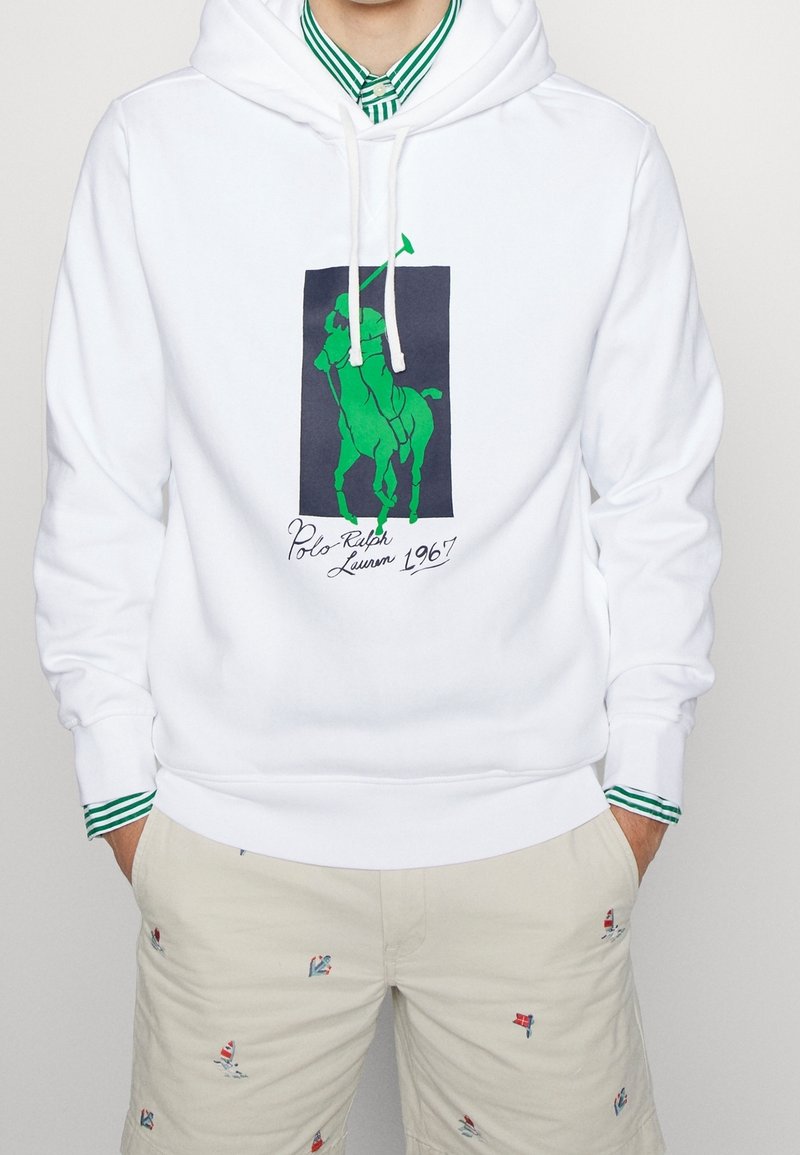 Mann trägt weißen Hoodie mit grünem Polo-Spieler-Logo und Text "1967", über grün gestreiftem Hemd, kombiniert mit beigen Shorts mit kleinen bunten Segelbootmotiven.