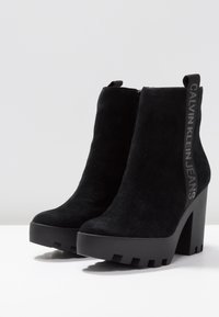 Mustat mokkanahkaiset nilkkurit, joissa on paksu korko, sivuvetoketju ja mustasta kankaasta valmistettu vetoketju, jossa on "Calvin Klein Jeans" -brändäys.