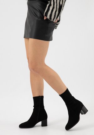 Jambes portant une mini-jupe en cuir noir, des bottines noires à talons carrés, et un pull à rayures zébrées avec les mains dans les poches.
