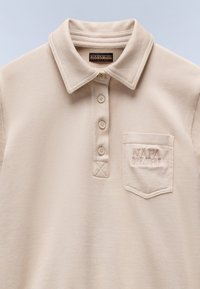Beige polo-paita, jossa on napitettu kaulus, ribbed-kaulus, teksturoitu kangas ja vasemmalla rinnalla tasku, jossa on brodeerattu logo.