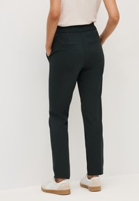 Pantalons élastiqués vert foncé avec poches latérales et détails de poche arrière, associés à des baskets blanches et beiges. Texture du tissu lisse.