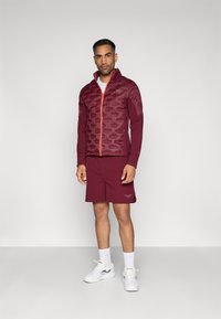 Bordový nafukovací kabát so zigzag vzorom a oranžovým zipsom, doplnený o zodpovedajúce šortky. Biely tenisky a ponožky dotvárajú celkový vzhľad.