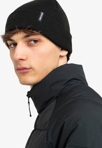 Berretto nero a maglia con un design a coste testurizzato; presenta un piccolo etichetta con il marchio. Il modello indossa una giacca nera con colletto alto.
