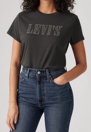 Vrouw draagt een zwart Levi’s t-shirt met gespijkerd logo en hooggetailleerde blauwe spijkerbroek, één hand in de zak tegen een effen achtergrond.