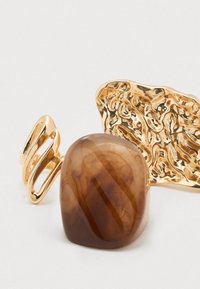 Design abstrait en ton or avec une pierre lisse marron foncé, présentant une texture marbrée et une forme arrondie, avec des détails organiques.