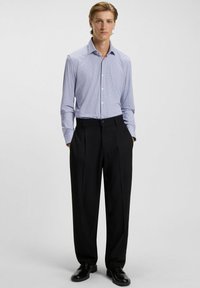 Hellblaues Hemd mit knöpfen und kleinem geometrischen Muster, kombiniert mit schwarzen, gefältelten Hosen und schwarzen Schuhen.