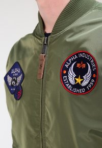 Giacca bomber verde oliva con collo a costine, zip frontale e due toppe ricamate: rotonda "Alpha Industries" e rettangolare con il logo "Alpha".