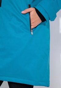 Teal buitenjack met een gladde textuur, voorzien van een zijzak met een zilveren rits en zwarte accenten bij de mouwen.