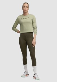 Felpa verde con il logo "HYROX", abbinata a leggings oliva. La modella indossa calze a righe bianche e nere e scarpe da ginnastica chiare.