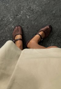 Brązowe buty typu Mary Jane z przezroczystą cholewką i pojedynczym paskiem, odsłaniające palce. Miękka, płaska podeszwa na teksturowanej powierzchni.