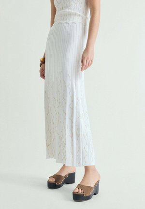 Mujer con una falda larga blanca acanalada con detalles dorados y sandalias mule marrones con plataforma sobre un fondo sencillo.