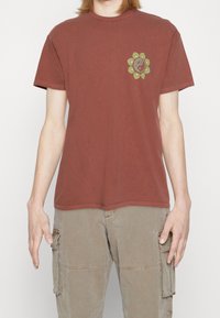 Röd bomullströja med rund hals, med en grön cirkulär blommig grafik på vänster bröstområde. Korta ärmar och avslappnad passform.