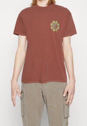 T-shirt en coton rouge à col rond, avec un graphique floral circulaire vert sur le côté gauche de la poitrine. Manches courtes et coupe décontractée.