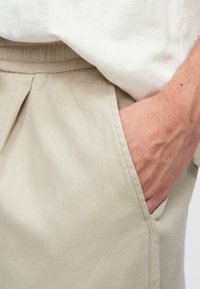 Pantalon en coton beige avec une coupe décontractée, doté d'une taille élastique et de poches latérales. Gros plan montrant la poche et la position de la main.