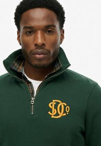 Sudadera de forro polar verde con cuello con cremallera, que presenta un bordado del logotipo en amarillo y un forro de franela a cuadros visible en el cuello.