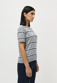 Woman with straight black hair wearing a gray short-sleeve sweater with navy and white stripes, and navy pants, standing against a plain background.