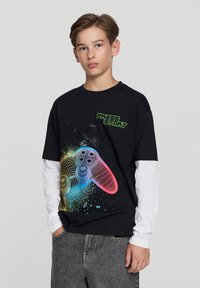 Czarny t-shirt z bawełny, z kolorową grafiką kontrolera gier oraz napisem "NACIŚNIJ START" w zielonym kolorze. Długie białe rękawy pod spodem.