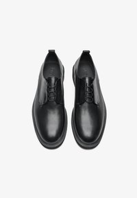 Zapatos de piel negra con un acabado suave y un diseño sin cordones. Presentan un lazo decorativo y una puntera redondeada. Suela plana con un dibujo mínimo.