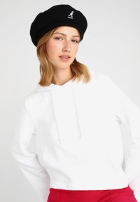 Sudadera blanca recortada con un bolsillo frontal y cordones, llevada con una boina negra con un logo, sobre un fondo neutro.