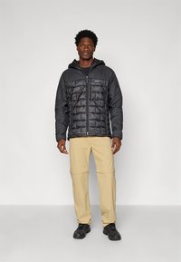 Juoda pūsta striukė su užtrauktuku priekyje ir gaubtu; turi violetinių akcentų ir siuvinėtą dizainą, derinama su smėlio spalvos kelnėmis ir juodais batais.