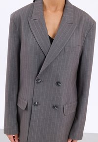 Blazer doppiopetto grigio a righe sottili con rever a punta, due tasche frontali e bottoni neri. Vestibilità sartoriale con spalle strutturate.