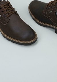Botas de hombre de cuero marrón oscuro con suelas negras resistentes, costuras en contraste y cordones redondos, contra un fondo gris claro.