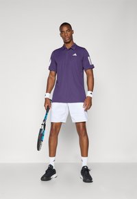 Violetti polo-paita valkoisilla yksityiskohdilla, valkoiset shortsit, mustat lenkkarit ja tennismaila. Paitassa ja käsivarsinauhoissa on Adidas-logo.