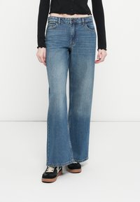 Lyseblå vide jeans laget av denim, med et klassisk fem-lomme design og slitte detaljer, kombinert med svarte joggesko.