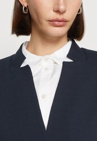 Blazer con colletto a navale e finitura texturizzata, indossato sopra una camicia bianca con bottoni di madreperla. Orecchini a cerchio in argento visibili.