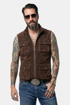 Mann mit Bart und Tätowierungen, der eine Sonnenbrille, eine braune Wildlederweste mit Taschen, geschichtete Halsketten, Ringe und eine große Gürtelschnalle über blauen Jeans trägt.