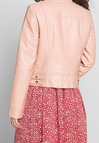 Světle růžová kožená bunda s prošívanými ramenními detaily, přiléhavý střih a dvojité zapínání v pase. Pod ní je viditeln dress s květinovým vzorem.