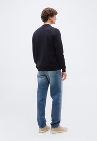 Νέος άνδρας που στέκεται με την πλάτη γυρισμένη, φοράει μαύρο μακρυμάνικο πουκάμισο, μπλε τζιν και μπεζ casual παπούτσια σε απλό λευκό φόντο.