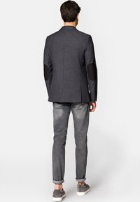 Męski ciemnoszary żakiet z brązowymi łatami na łokciach, dopasowany krój, w zestawie z jasnoszarymi spodniami i szarymi butami. Gładka tekstura materiału.